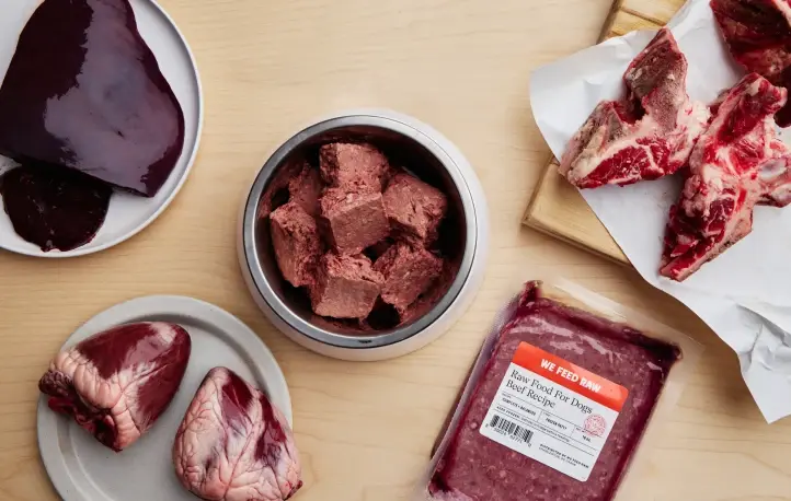 Assortment of raw dog food ingredients including organ meat, raw meat portions, bone-in cuts, and packaged beef blend from We Feed Raw on wooden surface.